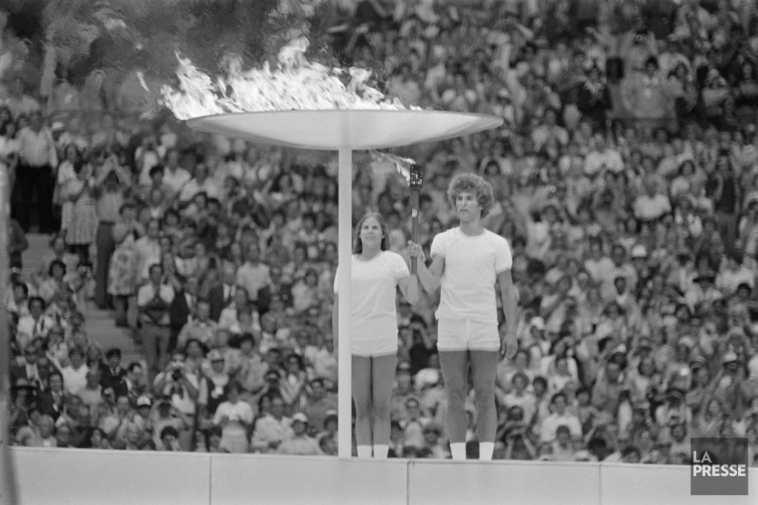 Photo noir et blanc de Sandra Henderson et Stéphane Préfontaine à côté de la flamme olympique lors de la cérémonie d’ouverture des Jeux olympiques de Montréal, le 17 juillet 1976