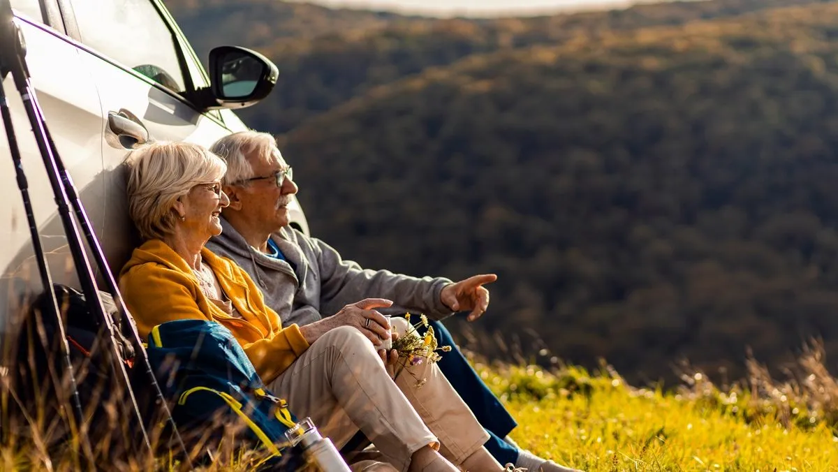 Un homme et une femme matures regardent un paysage de campagne, assis sur le sol, le dos contre leur voiture.