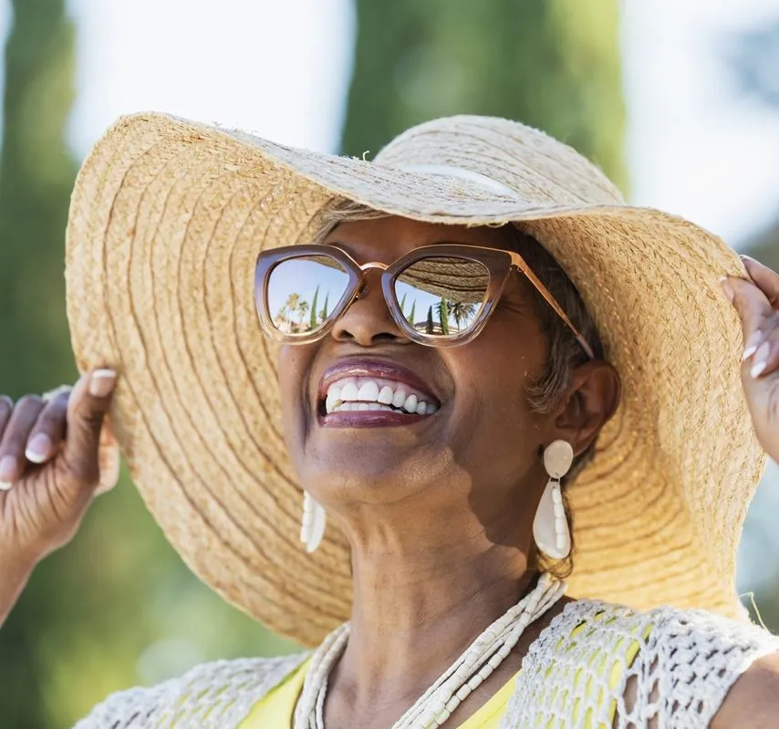 Une femme souriante et élégante porte un chapeau capeline et des lunettes de soleil.
