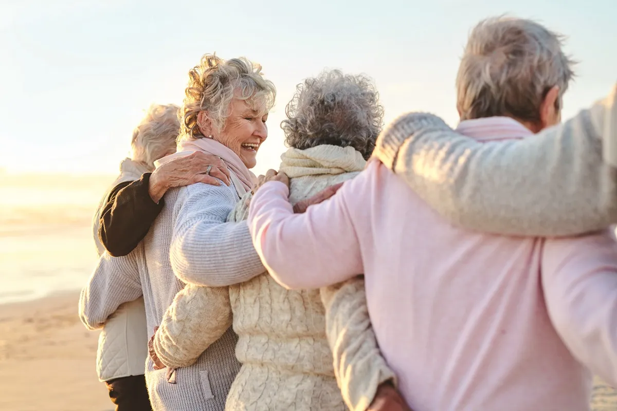 Groupe d'amies aux cheveux blancs marchant bras dessus bras dessous sur une plage, symbole d'une amitié de longue date.