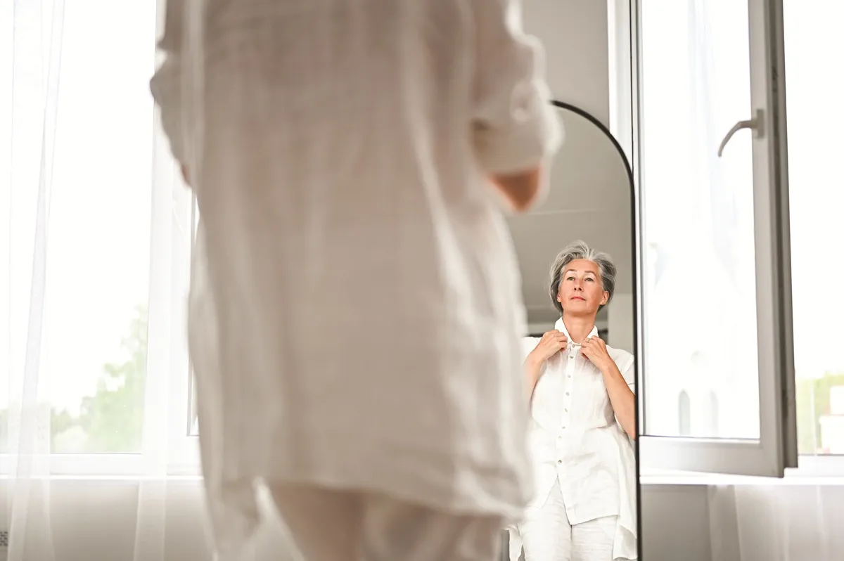 Femme aux cheveux argentés ajustant sa blouse devant un miroir dans une pièce lumineuse.