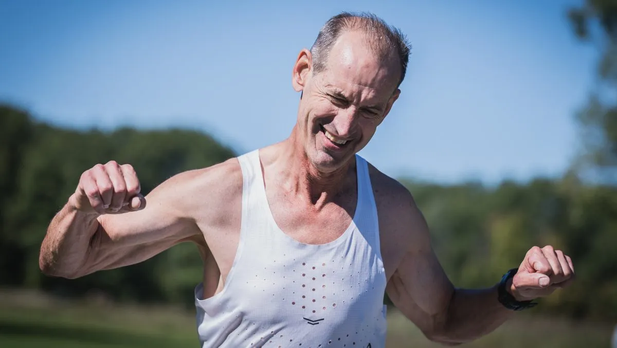 Participant aux Jeux FADOQ provinciaux levant les bras pendant une activité de course en plein air. Le sportif porte un débardeur blanc et une montre sportive, devant un décor de végétation sous un ciel dégagé.