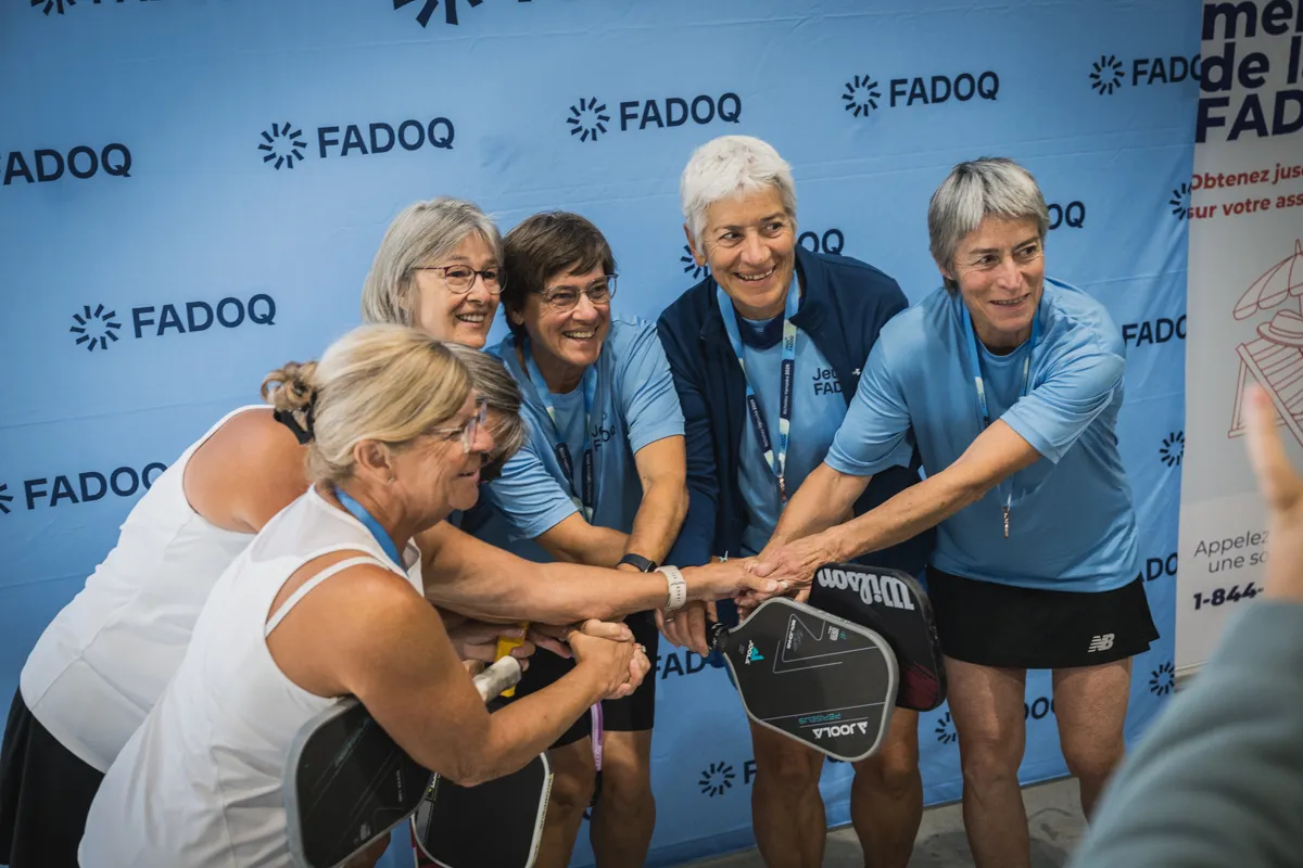 Des participantes aux Jeux FADOQ provinciaux se rassemblent devant un mur aux couleurs de la FADOQ pour une photo après la remise des médailles. Raquettes de pickleball en main, elles joignent leurs mains au centre dans un geste de solidarité et d’esprit sportif.
