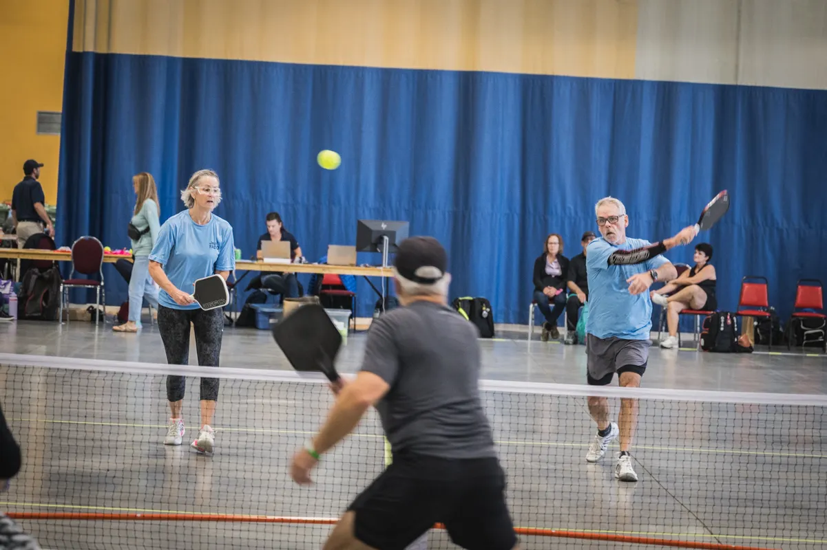 Des participants aux Jeux FADOQ provinciaux disputent une partie de pickleball dans un gymnase. Quatre joueurs sont visibles de part et d’autre du filet alors qu’une balle est en jeu devant des spectateurs et des officiels.