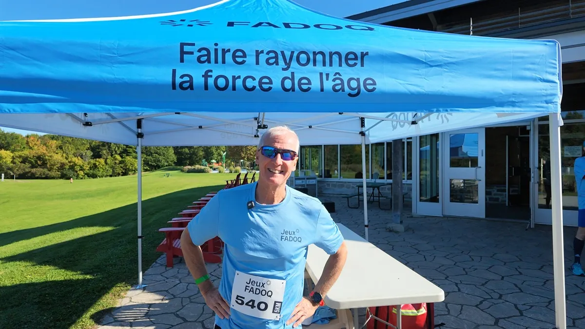 François Bernatchez porte son dossard des Jeux provinciaux de la FADOQ et pose devant une tente arborant le slogan « Faire rayonner la force de l’âge », à Saint-Hyacinthe, avant une course de 5 km.